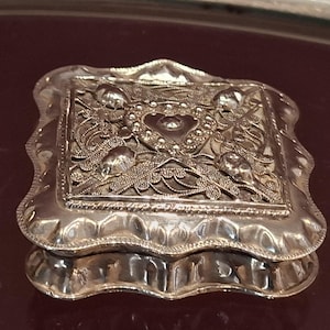 May include: A small, ornate silver box with a rectangular lid featuring a heart-shaped design and intricate floral patterns. The box has a scalloped edge and is set against a dark background.