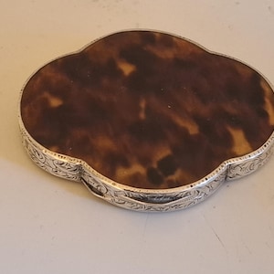 Op de afbeelding: Een vintage, sierlijk compactdoosje met een schildpad bovenkant en een zilverkleurig, gegraveerd metalen frame. De compact heeft een unieke, vierlobbige vorm en een gedetailleerd bloemmotief rond de randen.