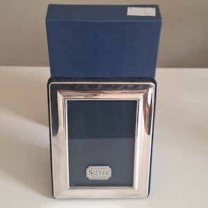 May include: A rectangular sterling silver picture frame with a polished metallic finish and a dark blue velvet backing. The frame features a small silver plaque at the bottom centre with the text Hallmark Sterling Silver Made In England.