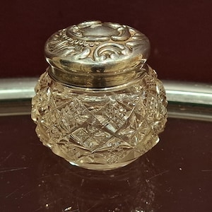 Pode incluir: Um pequeno frasco de vidro transparente com tampa prateada. O frasco tem um padrão de corte de diamante e a tampa é decorada com desenhos florais. O frasco está sobre uma superfície reflexiva.