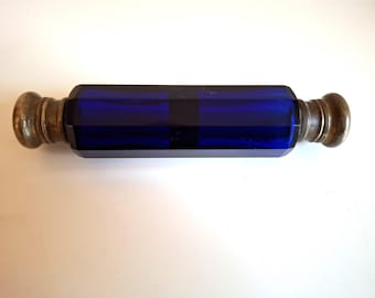 May include: A cobalt blue glass perfume bottle with faceted sides and metal end caps. The bottle has a cylindrical shape and is displayed horizontally against a white background. The metal caps have a tarnished appearance.