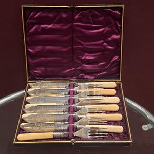 May include: A set of antique silverware, including knives and forks, presented in a black box with a purple velvet lining. The silverware features ornate silver handles and cream-colored grips. The box is open, revealing the arrangement of the utensils.