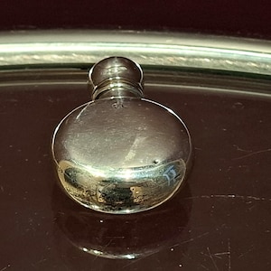 Könnte beinhalten: Kleine, runde, silberfarbene Parfümflasche mit abgerundeter Oberseite und kleinem, kugelförmigem Verschluss. Die Flasche reflektiert die Umgebung und erzeugt einen Spiegeleffekt. Sie steht auf einer dunklen, reflektierenden Oberfläche.