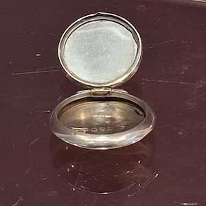 May include: Open silver compact with a hinged lid. The compact is round with a reflective surface inside the lid and a slightly convex base. The compact is on a dark surface.