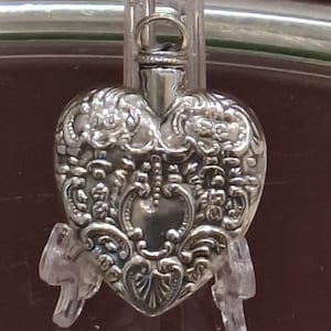 May include: A heart-shaped, silver-toned perfume bottle with intricate floral and scrollwork detailing. The bottle has a small loop at the top and is displayed on a clear stand. The design features a central heart shape and ornate embellishments.