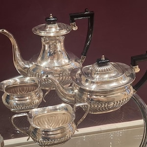 May include: A silver tea service set consisting of a tall teapot, a shorter teapot, a creamer, and a sugar bowl. Each piece features a polished silver finish with fluted detailing at the base and black handles and finials on the lids.