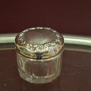 May include: A small, antique glass jar with a silver lid. The lid is round with a decorative floral pattern around the edge. The jar's body is clear, with a faceted design. The jar is sitting on a reflective surface.
