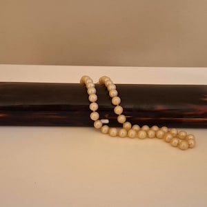 Pode incluir: Um estojo retangular vintage, castanho e âmbar, com um colar de pérolas drapeado sobre ele. O estojo tem um acabamento brilhante e bordas arredondadas. As pérolas são de cor creme.