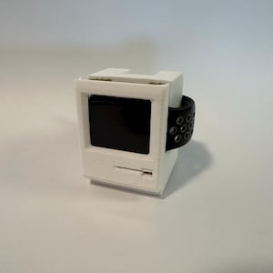Peut inclure: Un bracelet de montre imprimé en 3D blanc en forme d'ordinateur Macintosh d'ancienne génération. Le bracelet est doté d'un bracelet en silicone noir avec un design perforé.