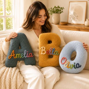 May include: Three plush, textured letter-shaped cushions in dark teal, mustard yellow, and light blue. The dark teal letter A features the name Amelia in rainbow-coloured script. The yellow letter B displays Bella, and the light blue O shows Olivia.