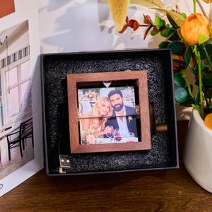 May include: A wooden rotating photo frame with a picture of a couple, presented in a black box. The frame is square and has a USB cable attached. The background includes a white vase with orange flowers and greenery.