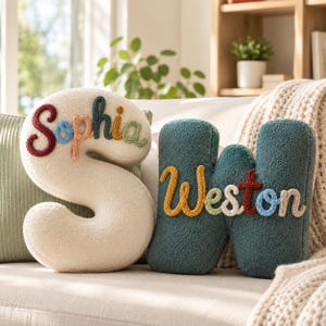 May include: Two plush letter-shaped cushions. The cream-coloured letter S features the name Sophia embroidered in multi-coloured yarn. The teal-coloured letter W features the name Weston embroidered in multi-coloured yarn. Both cushions have a textured surface.