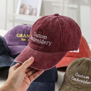 Op de afbeelding: Een bordeauxrode baseballcap met de tekst "Custom Embroidery" in het wit geborduurd. Op de achtergrond zijn andere baseballcaps in verschillende kleuren te zien, waaronder paars en olijfgroen.
