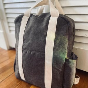 May include: A dark gray canvas backpack with white straps. The backpack has a zippered pocket on the front and a small pocket on the side.