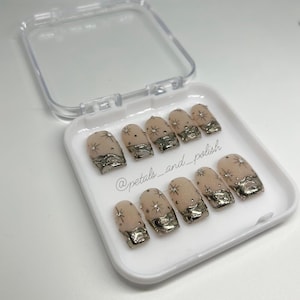 May include: A set of ten artificial nails in a white plastic case. The nails are nude-colored with silver metallic accents at the tips and small star-shaped embellishments. The text "@petals_and_polish" is visible.