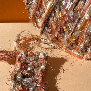 Peut inclure: Gros plan sur une pelote de fil texturé avec un petit échantillon. Le fil est un mélange de fibres orange, marron, argent et blanches. L'échantillon est rectangulaire et fait du même fil. Le fil semble être pour l'artisanat.