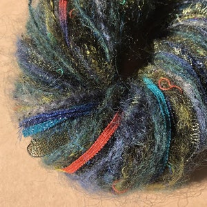 May include: A close-up of a ball of yarn with a variety of colors, including green, blue, gold, and red. The yarn is made of a soft, fluffy material and has a slightly fuzzy texture.