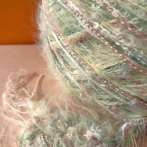 May include: A skein of yarn with a mix of white, green, and pink fibers. The yarn is fluffy and has a soft, fuzzy texture.