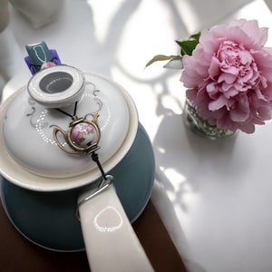 Op de afbeelding: Een turquoise en witte porseleinen theepot met een bloemenmotief en deksel. Een kleine theepotbedel hangt aan het deksel. Een roze pioenroos in een glazen vaas staat ernaast.