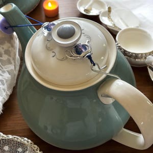 Op de afbeelding: Een teal keramische theepot met een witte handgreep en deksel. Het deksel heeft een zilver en blauw decoratief element. Een kleine kaars en theekopjes staan op de achtergrond, wat wijst op een theeset.