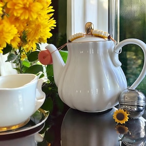 Pode incluir: Uma chaleira de cerâmica branca com tampa e alça douradas, uma chávena de chá a condizer e um infusor de chá em aço inoxidável. Um pequeno girassol decorativo e um ramo de flores amarelas também estão presentes. A chaleira tem uma capacidade de aproximadamente 1,2 litros.