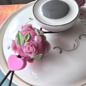 Op de afbeelding: Een wit keramisch theepotdeksel met een grijze knop en een roze rozenversiering. De roos is omgeven door groene bladeren en heeft een roze hartvormige bedel. Het theepotdeksel heeft een decoratief wervelontwerp.