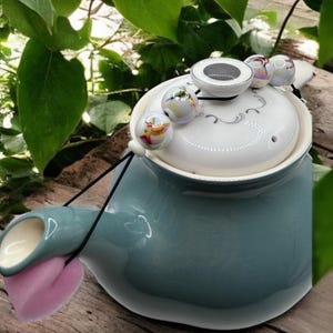 Könnte beinhalten: Eine türkisfarbene Keramik-Teekanne mit weißem Deckel mit dekorativem Design. Der Deckel ist mit irisierenden Glasperlen und einem kleinen, runden, weißen Keramikstück verziert. Ein rosa Filzstück ist am Ausguss befestigt.