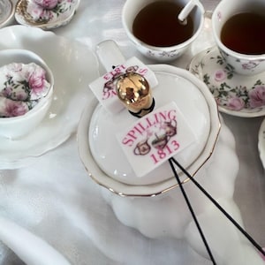 May include: White teapot with a gold trim and two black sticks sticking out of the lid. The teapot has two labels on it, one that says "E181" and the other that says "SPILLING The Tea 1813".