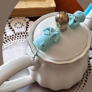 Puede incluir: Tetera de porcelana blanca con borde dorado, pomo dorado y dos sujetadores de bolsitas de té en forma de uniforme médico azul claro. La tetera está sobre un mantel de encaje blanco.