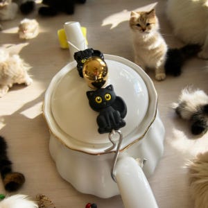 May include: A white teapot with a gold trim and a black cat charm hanging from the spout. The charm has large yellow eyes. There are several kittens in the background.