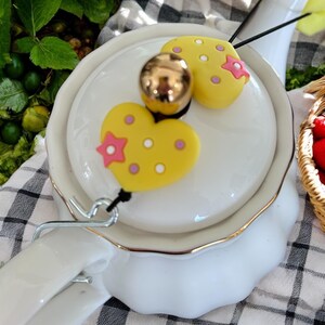 May include: White ceramic teapot with a gold trim and a lid featuring two yellow heart-shaped ornaments with pink polka dots and a pink star. The ornaments are attached to the lid with a black string.