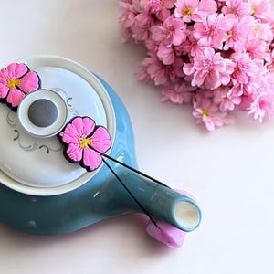 Può includere: Una teiera blu-verde con un coperchio bianco e due accenti di fiori rosa. La teiera ha un manico in corda nera e un beccuccio bianco.