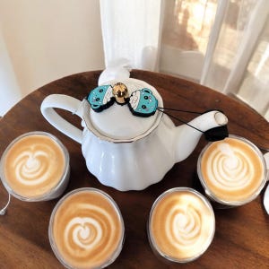 Könnte beinhalten: Eine weiße Teekanne mit goldener Verzierung und zwei blauen und weißen Cartoon-Teetassenanhängern am Henkel. Vier Tassen Kaffee mit Latte Art stehen auf einem Holztisch.