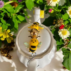 Könnte beinhalten: Drei gelbe und schwarze Bienen-Anhänger mit weißen Flügeln und lächelnden Gesichtern auf einer weißen Teekanne. Die Anhänger sind an einer goldenen Kette befestigt.