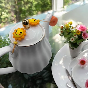 Könnte beinhalten: Eine weiße Keramik-Teekanne mit goldenem Rand, verziert mit gelben sonnenförmigen Anhängern. Die Teekanne ist Teil eines Teeservices, mit einer kleinen Blumenvase und einem Teller im Hintergrund. Die Teekanne hat ein einzigartiges, gerüschtes Design.