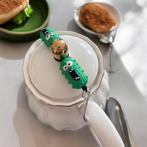 Op de afbeelding: Een novelty thee-infuser met twee groene augurk-vormige bedels met zwarte gezichten en een goudkleurige bol. De infuser is bevestigd aan een witte keramische theepot met een gouden rand. De achtergrond bevat een groene kom en een klein bord.