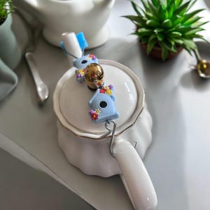 Könnte beinhalten: Eine weiße Teekanne mit einem blauen und floralen Tee-Infuser. Der Infuser ist in Form eines Vogelhauses mit einem goldenen Akzent.