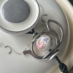 Può includere: Un ciondolo a forma di teiera argentato con una perla di porcellana bianca decorata con un motivo floreale rosa e viola.