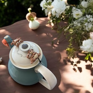 Puede incluir: Una tetera azul y blanca con una tapa de cerámica y un infusor de té. El infusor está decorado con tres pequeños osos de cerámica marrones.