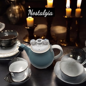 May include: A blue and white teapot with a decorative lid, featuring two crystal-like embellishments. The word "Nostalgia" is written in black script above the teapot. The scene includes lit candles and teacups, suggesting a tea party setting.