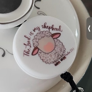 Könnte beinhalten: Runde weiße Plakette mit einer Illustration eines rosa und weißen Schafs und dem Text "The Lord is my shepherd, Psalm 23:1".