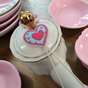 May include: A white porcelain teapot with a heart-shaped lid and gold trim. The lid features a pink and blue heart design with white polka dots. The teapot is part of a tea set with matching cups and saucers.