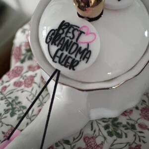 May include: White ceramic teapot with a gold trim and a black and pink embroidered patch that says "Best Grandma Ever".
