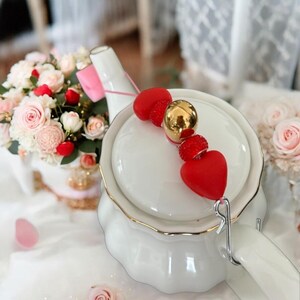 Peut inclure: Théière blanche avec une bordure dorée et un charme en forme de cœur rouge attaché au bec verseur. Le charme est fait de perles rouges et d'une perle dorée.