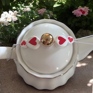 Op de afbeelding: Witte keramische theepot met een gouden rand en deksel. Een decoratieve streng witte kralen met rode harten en een gouden kraal siert het deksel. De achtergrond toont groen gebladerte en roze bloemen.