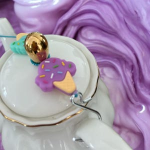 Può includere: Una teiera bianca con un bordo dorato e un ciondolo a forma di gelato viola, blu e giallo attaccato al beccuccio.