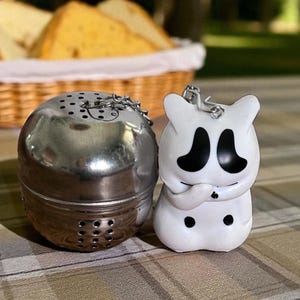 May include: A stainless steel tea infuser with a chain next to a white ghost-shaped keyring with black eyes and a mouth. The tea infuser is round with small holes. The keyring has a silver metal loop.