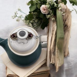 Könnte beinhalten: Eine teal-farbene und weiße Porzellan-Teekanne mit einem dekorativen Deckel mit zwei lila Edelsteinen. Die Teekanne steht auf einem Stapel alter Bücher und Papiere, mit einem Blumenstrauß und Bändern im Hintergrund.