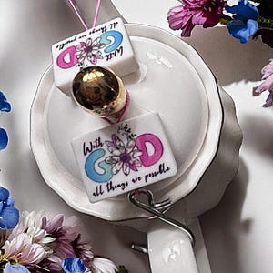 Op de afbeelding: Een witte theepot met een gouden rand en een bloemenmotief. De theepot heeft een deksel met een handvat en een tuit. Twee kleine witte keramische tegels met de woorden "Met God is alles mogelijk" zijn met een touwtje aan de theepot bevestigd.