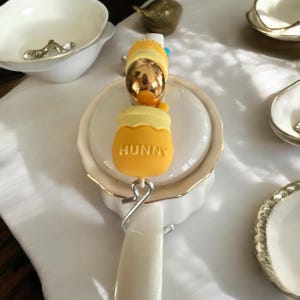 Op de afbeelding: Witte theepot met een gouden rand en een Winnie de Poeh-thema honingpot charme. De charme is geel en zegt "Hunny" erop.